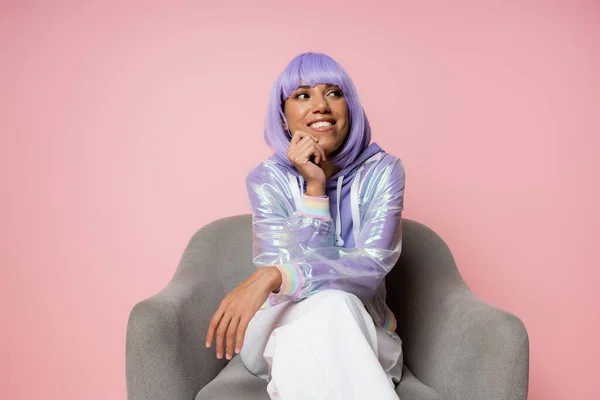 Mujer afroamericana feliz en peluca púrpura sonriendo mientras está sentado en sillón gris aislado en rosa - foto de stock