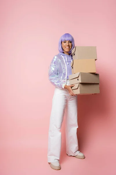 Comprimento total de mulher americana africana feliz em roxo peruca segurando caixas de papelão em rosa — Fotografia de Stock