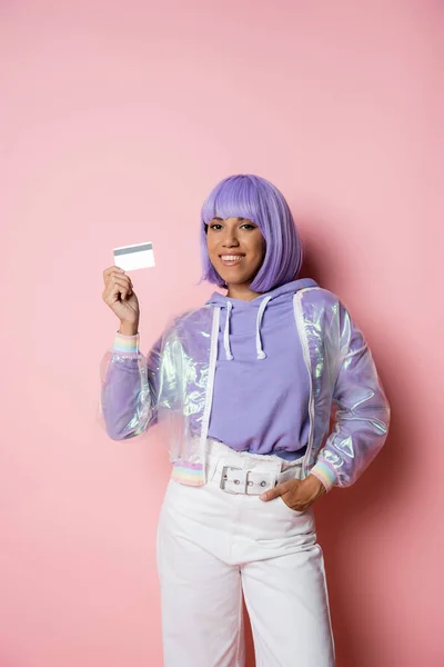 Femme américaine africaine joyeuse avec les cheveux violets tenant la carte de crédit et posant avec la main dans la poche sur rose — Photo de stock