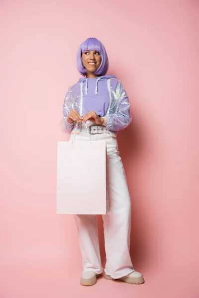 Pleine longueur de femme africaine américaine gaie avec les cheveux violets tenant sac à provisions sur rose — Photo de stock