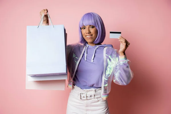Alegre mujer afroamericana en chaqueta transparente sosteniendo bolsas de compras y tarjeta de crédito en rosa - foto de stock