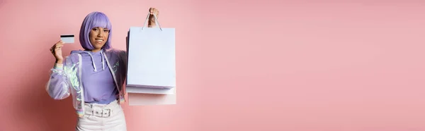 Mulher americana africana alegre em jaqueta transparente segurando sacos de compras e cartão de crédito em rosa, banner — Fotografia de Stock