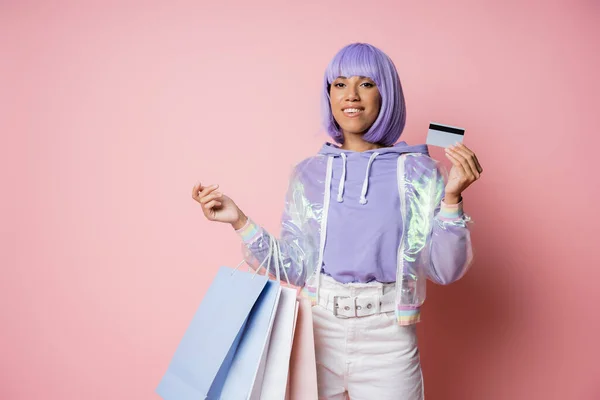 Alegre mujer afroamericana en chaqueta transparente sosteniendo bolsas de compras y tarjeta de crédito en rosa - foto de stock