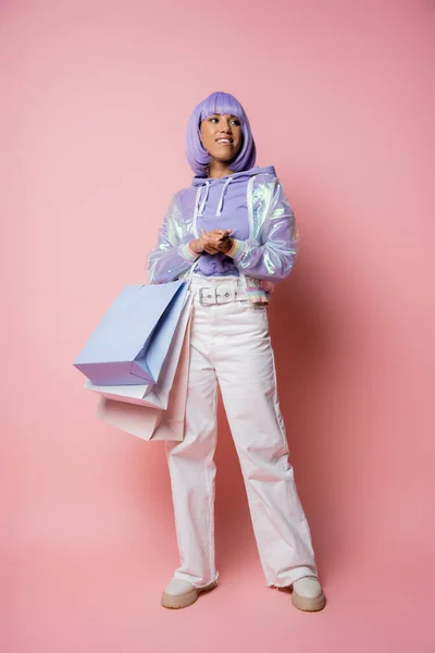 Longitud completa de la alegre mujer afroamericana en chaqueta transparente de pie con bolsas de compras en rosa - foto de stock