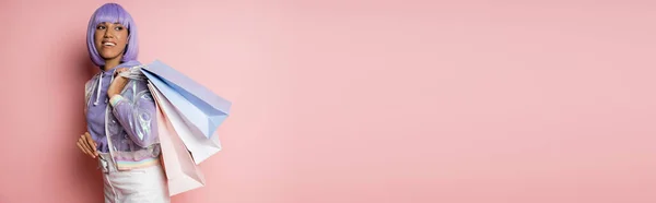 Alegre mujer afroamericana con el pelo morado de pie en chaqueta transparente con bolsas de compras en rosa, bandera - foto de stock