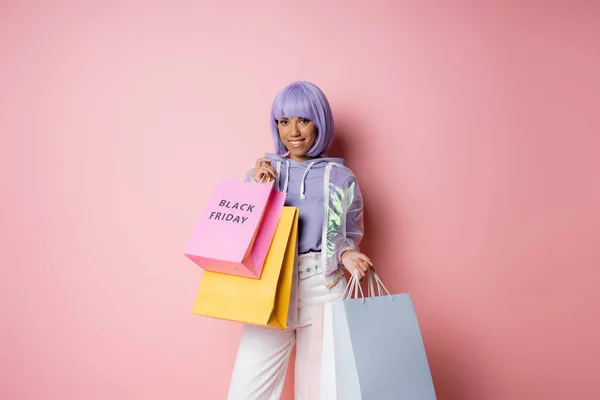 Positive femme afro-américaine aux cheveux violets tenant des sacs à provisions avec lettrage noir vendredi sur rose — Photo de stock