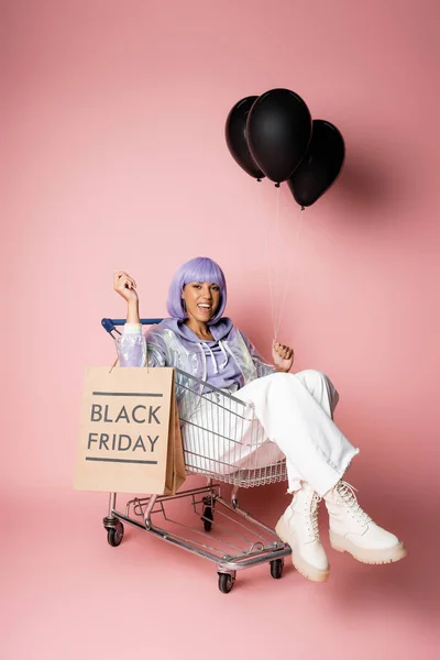 Pleine longueur de femme africaine américaine gaie tenant sac à provisions avec lettrage noir vendredi tout en étant assis dans le panier sur rose — Photo de stock