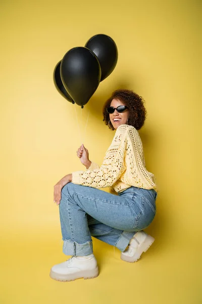 Volle Länge der fröhlichen afrikanisch-amerikanischen Frau mit Sonnenbrille hält schwarze Luftballons, während sie auf gelb sitzt — Stockfoto