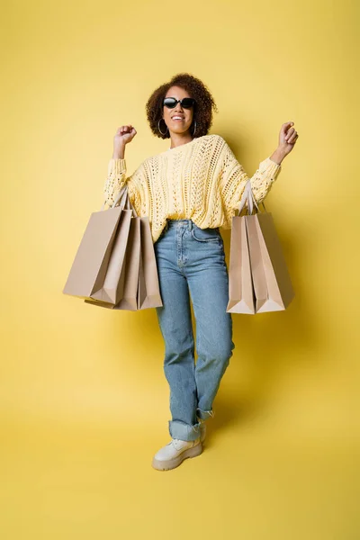 Volle Länge der fröhlichen afrikanisch-amerikanischen Frau in Sonnenbrille mit Einkaufstaschen auf gelb — Stockfoto