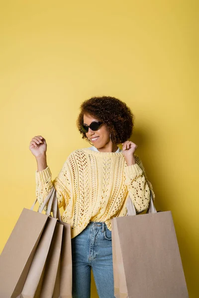 Mulher americana africana alegre em óculos de sol segurando sacos de compras em amarelo — Fotografia de Stock