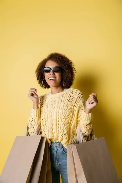 Веселая африканская американка в стильных солнцезащитных очках, держащая сумки с покупками на желтом — стоковое фото