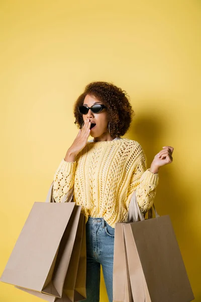 Amazed african american woman in stylish sunglasses holding shopping bags on yellow — Stock Photo