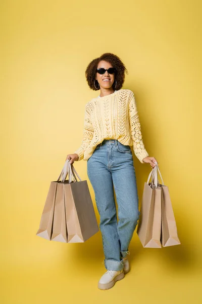 Full length of happy african american woman in sunglasses holding shopping bags on yellow — Stock Photo