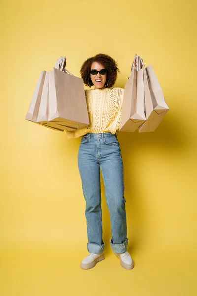 Pleine longueur de femme afro-américaine positive dans des lunettes de soleil tenant des sacs à provisions sur jaune — Photo de stock