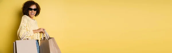Allegra donna afroamericana in eleganti occhiali da sole che tiene borse della spesa su giallo, banner — Foto stock