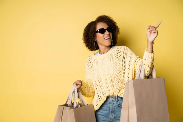 Mulher americana africana feliz em óculos de sol elegantes segurando sacos de compras e apontando com o dedo no amarelo — Fotografia de Stock