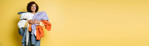 Счастливая африканская американка с кучей разноцветной одежды на желтом фоне, баннер — стоковое фото