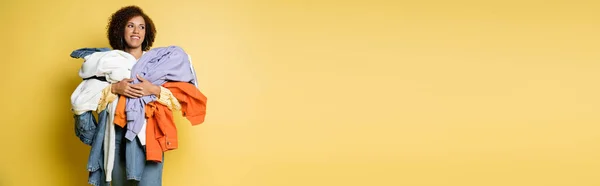 Mujer afroamericana feliz sosteniendo pila de ropa colorida sobre fondo amarillo, bandera — Stock Photo