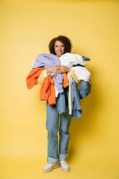 Longitud completa de la mujer afroamericana feliz sosteniendo pila de ropa colorida en amarillo - foto de stock