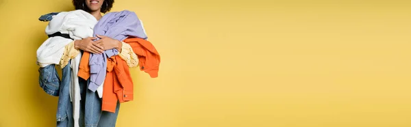 Vista recortada de la mujer afroamericana feliz sosteniendo pila de ropa colorida en amarillo, bandera - foto de stock