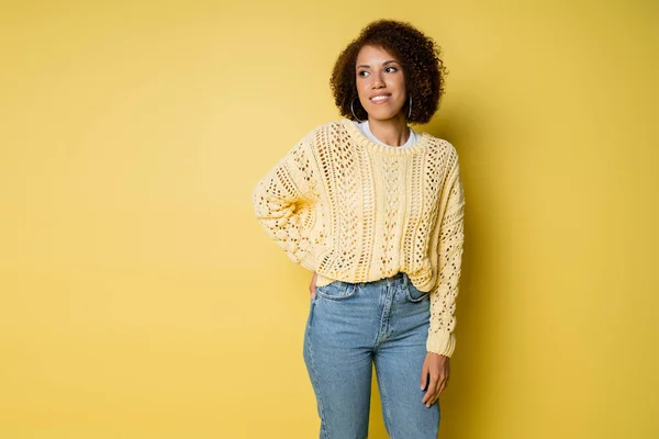 Allegra e giovane donna afroamericana in maglia maglione posa con mano sul fianco su giallo — Foto stock