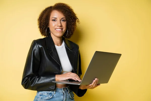 Mulher americana africana feliz e jovem em jaqueta de couro usando laptop em amarelo — Fotografia de Stock