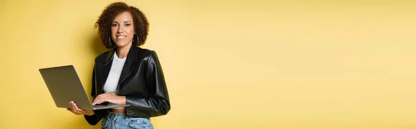 Mujer americana africana feliz y joven en chaqueta de cuero usando el ordenador portátil en amarillo, bandera - foto de stock