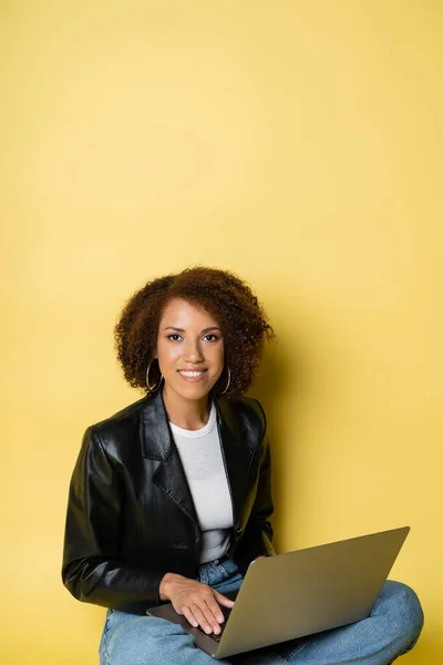 Fröhliche und junge afrikanisch-amerikanische Frau in Lederjacke mit Laptop auf gelb — Stockfoto