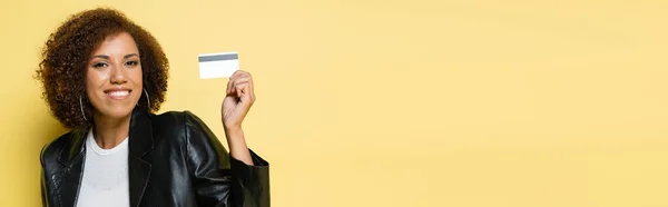 Freudige afrikanisch-amerikanische Frau in Lederjacke mit Kreditkarte auf gelbem Banner — Stockfoto