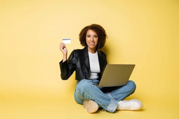 Alegre mujer afroamericana en chaqueta de cuero con tarjeta de crédito mientras está sentado con el ordenador portátil en amarillo - foto de stock