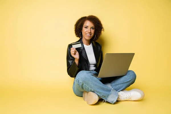 Mujer americana africana alegre en chaqueta de cuero que sostiene la tarjeta de crédito mientras que se sienta con el ordenador portátil en amarillo - foto de stock
