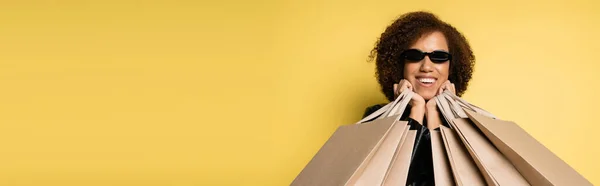 Femme afro-américaine gaie dans des lunettes de soleil tenant des sacs à provisions le vendredi noir sur jaune, bannière — Photo de stock