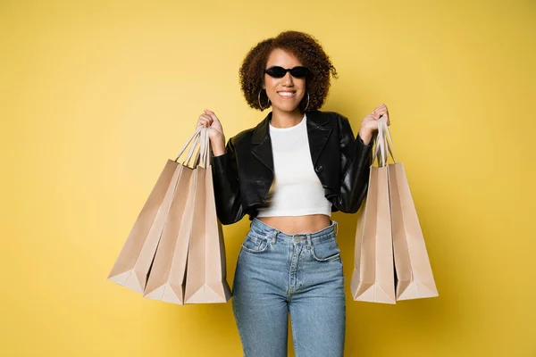 Positive african american woman in sunglasses and leather jacket holding shopping bags on yellow — Stock Photo