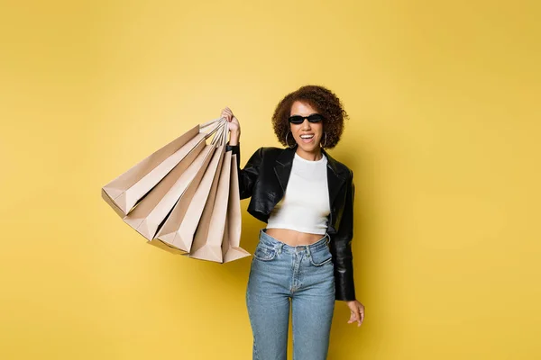 Excited african american woman in stylish sunglasses and leather jacket holding shopping bags on yellow — Stock Photo