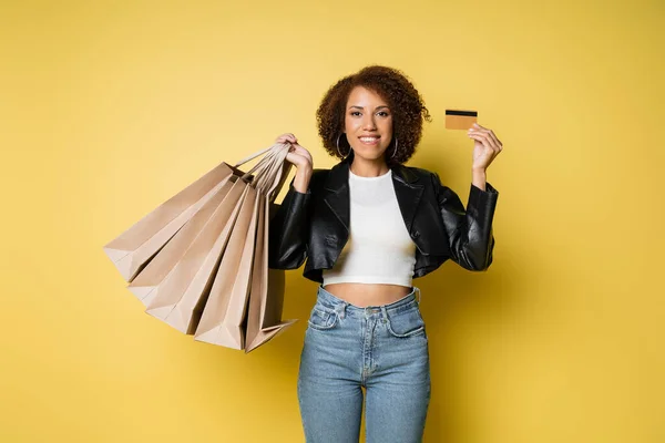 Femme afro-américaine gaie en veste en cuir élégant tenant des sacs à provisions et carte de crédit sur jaune — Photo de stock
