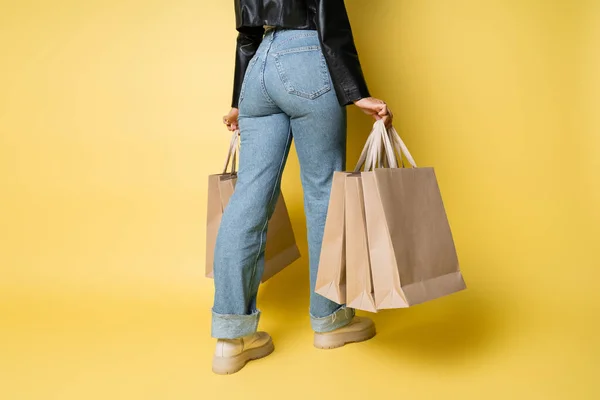Cropped view of african american woman in stylish leather jacket and denim jeans holding shopping bags on yellow — Stock Photo