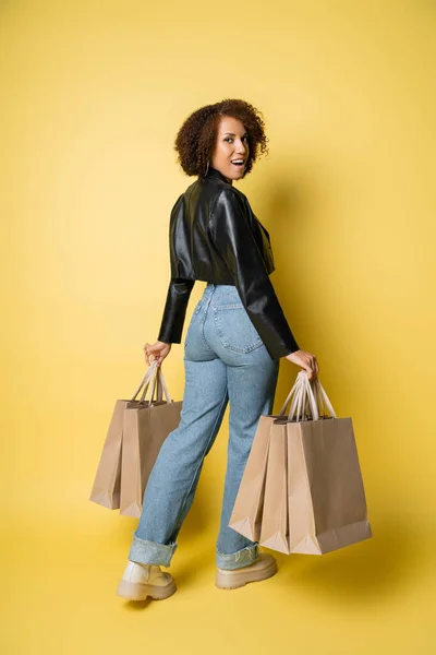 Full length of positive african american woman in stylish leather jacket and denim jeans holding shopping bags on yellow — Stock Photo