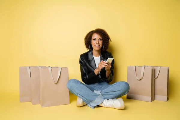 Piena lunghezza di felice donna afroamericana in giacca di pelle e jeans utilizzando smartphone vicino shopping bags su giallo — Foto stock