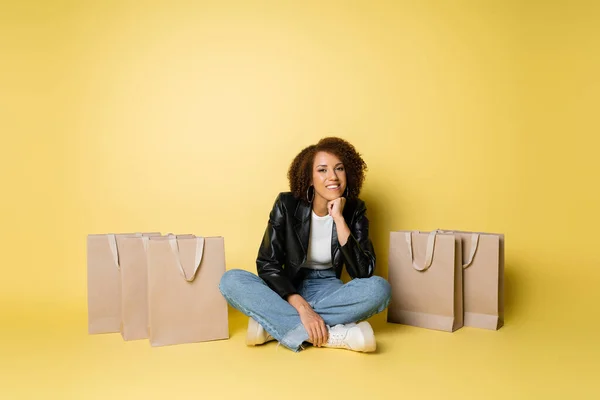 Piena lunghezza di felice donna africana americana in giacca di pelle e jeans seduto vicino a borse della spesa su giallo — Foto stock