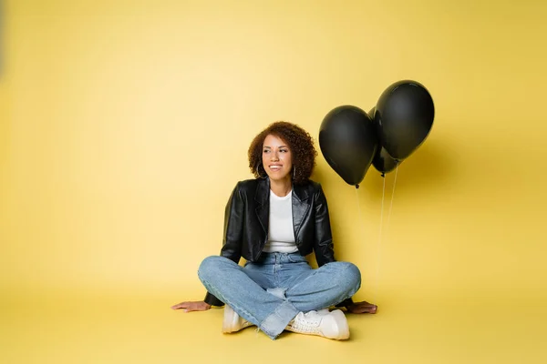 Full length of smiling african american woman in leather jacket and jeans sitting near black balloons on yellow — Stock Photo