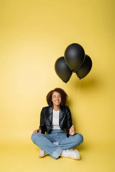 Full length of happy african american woman in leather jacket and jeans sitting near black balloons on yellow — Stock Photo