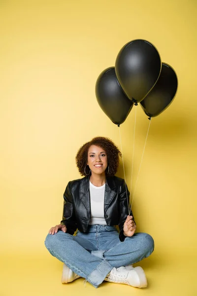 Pleine longueur de femme afro-américaine heureuse en veste en cuir et jeans assis avec des ballons noirs sur jaune — Photo de stock