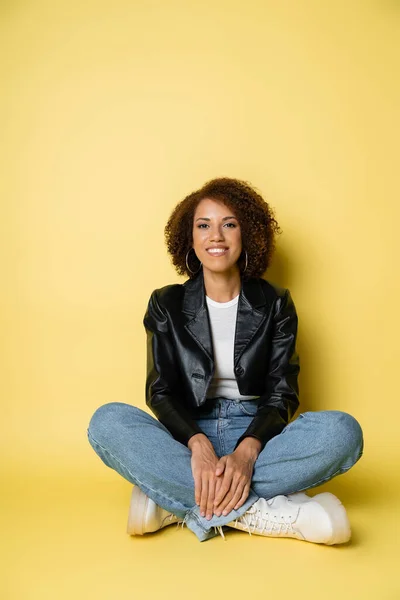 Full length of pleased african american woman in leather jacket and jeans sitting with crossed legs on yellow — Stock Photo