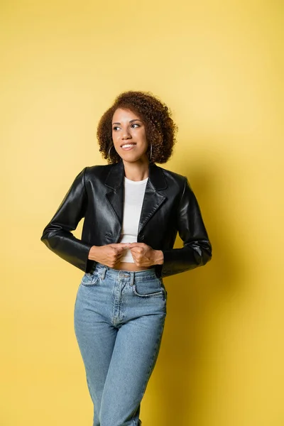 Mulher americana africana satisfeito em jaqueta de couro elegante e jeans posando no amarelo — Fotografia de Stock