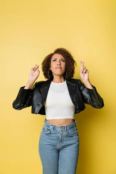 Curly african american woman in stylish leather jacket and jeans posing with crossed fingers on yellow — Stock Photo