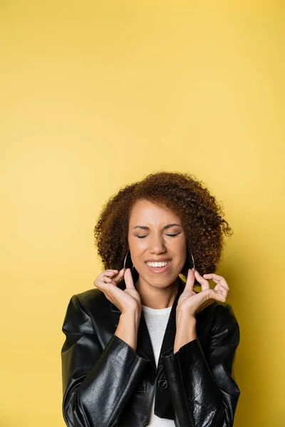 Pleased african american woman in stylish leather jacket touching hoop earrings on yellow — Stock Photo