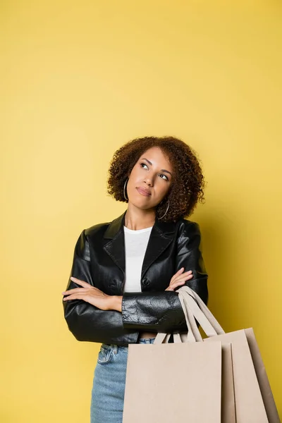 Mujer afroamericana pensativa en elegante chaqueta de cuero con bolsas de compras con compras en amarillo - foto de stock