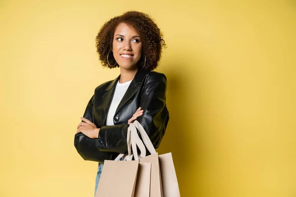 Mujer afroamericana feliz en elegante chaqueta de cuero sosteniendo bolsas de compras y de pie con los brazos cruzados en amarillo - foto de stock