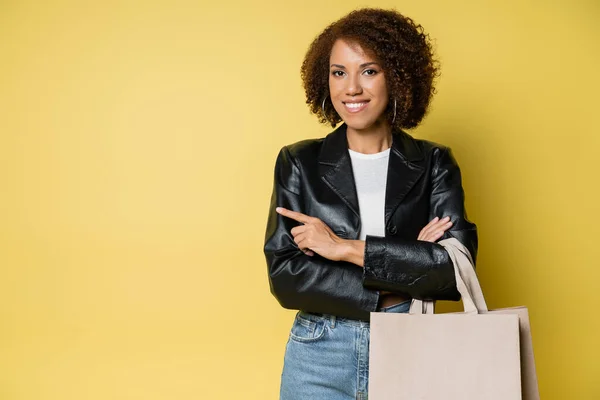 Mulher americana africana alegre em jaqueta de couro elegante segurando sacos de compras com compras e apontando com o dedo no amarelo — Fotografia de Stock
