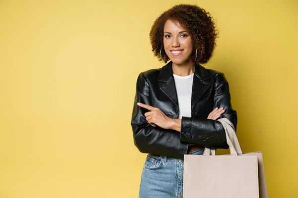 Cheerful african american woman in stylish leather jacket holding shopping bags with purchases and pointing with finger on yellow — Stock Photo